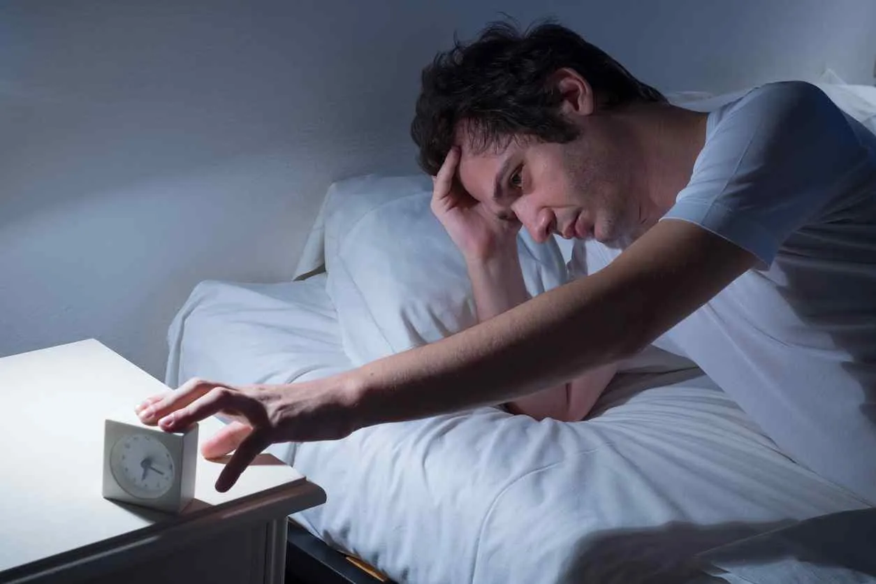 Man awake at night in bed reaching for an alarm clock, a common picture of disrupted sleep